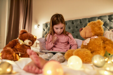 A cute small girl wearing pyjamas sitting alone on a huge bed barefoot scrolling photos in a tablet with her teddybears around the bed