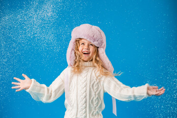 little blonde girl in winter clothes blows snow off her hands. blue background, space for text