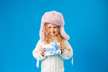 Obraz premium Cute little blonde girl in a pink hat and white knitted sweater smiles and holds a gift box on a blue background. Space for text
