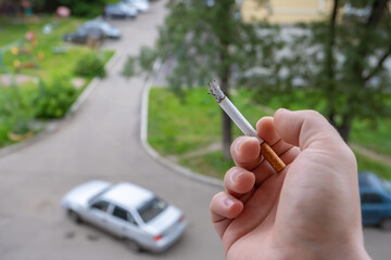 Smoking cigarette with ash and filter in the hand of a smoker who smokes in the window of the house and watches the passing cars in the yard