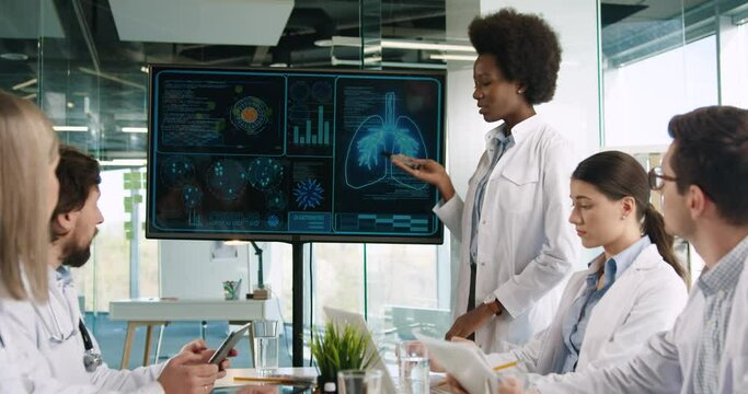 Mixed-races male and female doctors sitting on meeting in hospital office looking at digital screen. African American medical worker speaking about pulmon disease covid-19 coronavirus pandemic