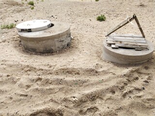 Sand, concrete, iron and wood.
Sandy area with sparse green grass. There are two concrete wells shafts, one covered with a concrete slab and the other with an iron hatch.