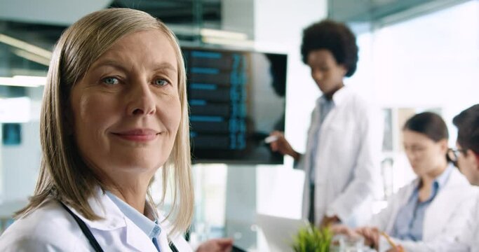 Close up portrait of happy senior female doctor turning to camera and smiling with mixed-races male and female doctors discussing pulmon disease covid-19 on meeting. Coronavirus pandemic concept