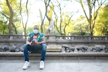 Obraz premium A young black man with a mask in the covid-19 pandemic season for protection. African American man with cell phone. Portrait of a young african man with a face mask in park.
