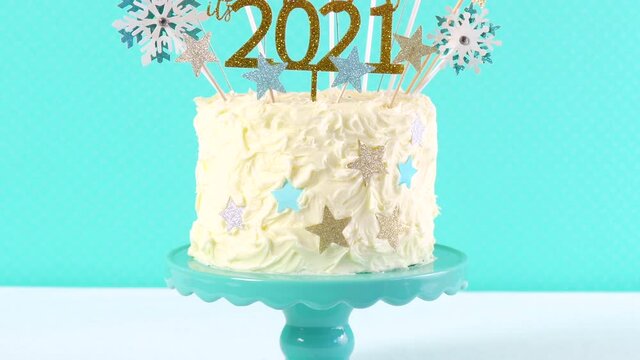 Happy New Year's Eve celebration cake on cake stand in blue white and gold theme decorated with stars and humorous, Thank God It's 2021, cake decoration with burning sparkler. Close up panning up.
