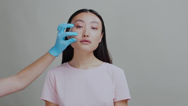 face portrait of healthy attractive chinese woman standing on grey background. Cosmetologist hands checking skin njecting hyaluronic acid into cheecks. beauty center.