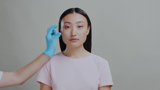 Portrait of chinese female face isolatedon grey background. Cosmetologist hands injecting hyaluronic acid cheeck injection for face anti-aging skincare procedure.