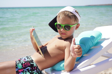 funny boy uses tablet on the beach. A child is sitting in a sun lounger on the beach against the background of the azure sea with a tablet in his hands.