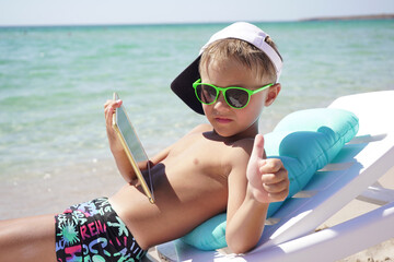funny boy uses tablet on the beach. A child is sitting in a sun lounger on the beach against the background of the azure sea with a tablet in his hands. © Katy_81