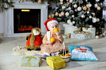 Beautiful girl in a red Christmas hat near the Christmas tree. She is sitting on the carpet with toys. A girl holds fruit in her hands. Next to the fireplace. The concept of the celebration
