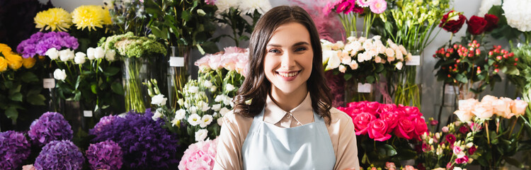 Happy brunette florist looking at camera with racks of flowers on background, banner