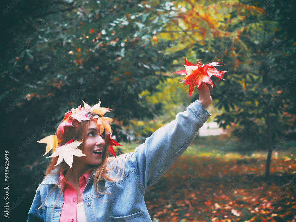 Wall mural A girl with a head decorated with red maple leaves holds a bouquet of autumn leaves in a park in her raised hand