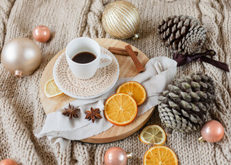 Fototapeta na wymiar cup of coffee on wooden round tray on knitted beige blanket background with Christmas decorations around it: Christmas ornaments, pine cones, ribbons, dried oranges, cinnamon and anise