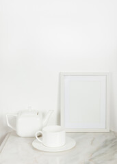 Fototapeta na wymiar Blank white photo frame on marble surface against white wall, near a white porcelain teapot and a. white tea cup 