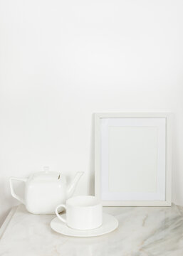 Blank white photo frame on marble surface against white wall, near a white porcelain teapot and a. white tea cup © irina_lazutina