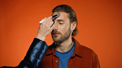 makeup artist applying face powder on young bearded man on orange