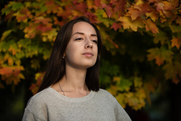A young girl stands against the background of an autumn maple tree. He smiles slightly and looks into the distance.