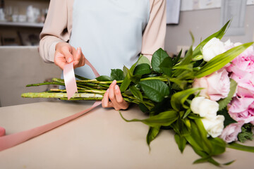 Cropped view of female florist with decorative ribbon tying stalks of fresh bouquet in flower shop on blurred background