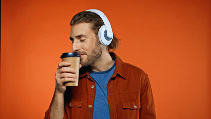 bearded man in wireless headphones smelling coffee to go in paper cup and listening music on orange
