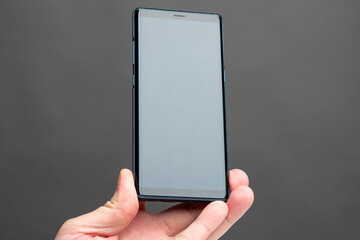 Person holds a smartphone in an upright position. Hand with a mobile phone in the center of the photo. Smartphone with a blank screen on gray background. Smartphone with space for text on the screen.