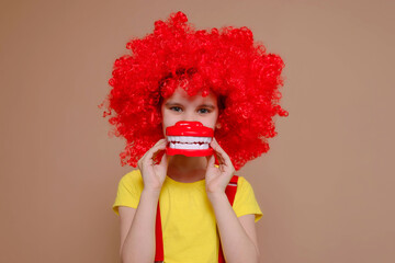 Funny clown girl child in a red wig and with a nose holding a model of a tooth and brush isolated against the background of a Set Sail Champagne flower. Kid teeth hygiene, teeth cleaning concept.