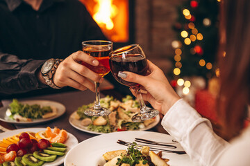 hands with glasses clinking against the background of Christmas tree lights and bonfires from a home fireplace over a table with delicious dishes
