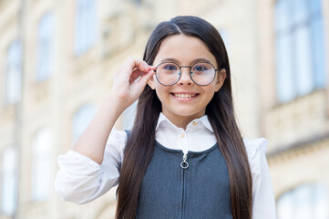 Happy girl child wearing school uniform fix eyeglasses in fashion frame to see clearly outdoors, opticians