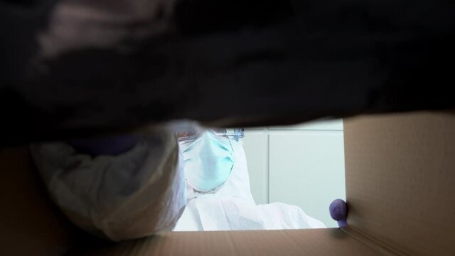 Doctor in protective suit and medical mask opening cardbox and stretching hand inside of enclosure during Covid19. Male nurse on PPE looking inside carton box at interior clinic. Coronavirus concept