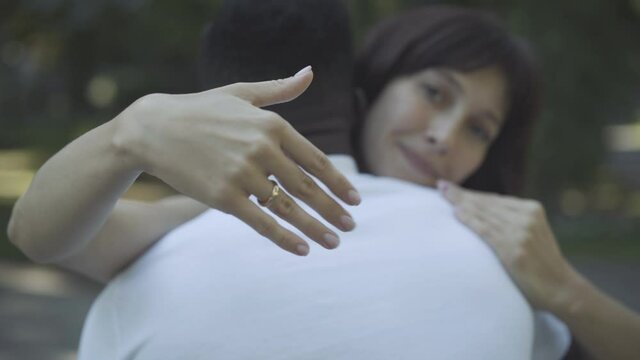 Close-up of female Caucasian hand with engagement ring, blurred woman hugging African American man at the background. Happy loving woman bragging jewelry outdoors. Interracial marriage concept.