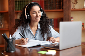 Woman sitting at desk, using laptop and writing in notebook