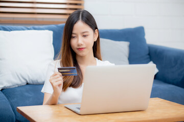 Young asian woman sit on sofa using laptop computer shopping online with credit card buying to internet, happy girl payment with e-business on couch, purchase and payment, business concept.