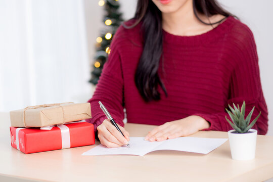 Closeup Hand Of Young Asian Woman Writing Postcard In Christmas Day At Home, Eve And Celebrate, Female Writing Message On Greeting Card With Giving Gift Box In Holiday, Congratulation And Celebration.