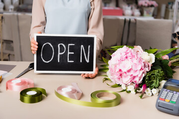 Cropped view of florist holding chalkboard with open lettering near bouquet on desk on blurred background