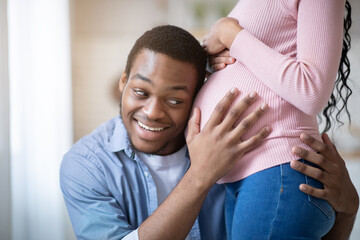 Fototapeta premium Caring black guy putting his ear on girlfriend's pregnant tummy, listening to baby's heartbeat at home