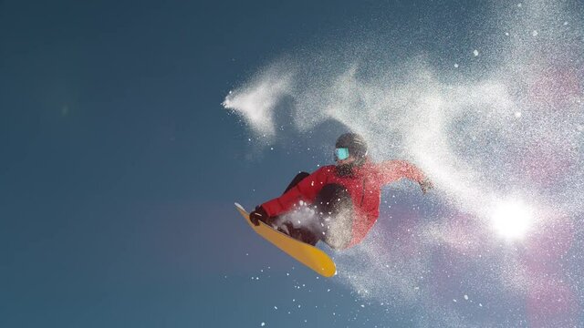 SLOW MOTION TIME WARP, LENS FLARE, BOTTOM UP: Fearless snowboarder does a spin trick while riding off a big air kicker. Snowflakes sparkle in the sunshine as male tourist does snowboarding tricks.