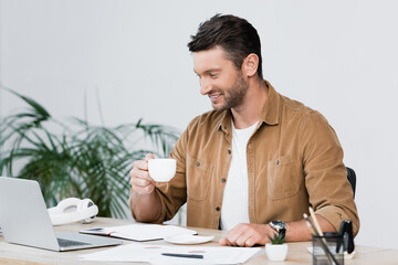 Cheerful businessman with coffee cup looking at laptop while sitting at workplace with blurred plant on background