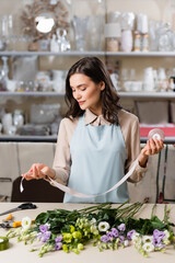 florist in apron holding decorative ribbon near eustoma flowers on table near rack with vases on blurred foreground