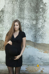 Beautiful young woman in a black dress and a black hat against the old wall