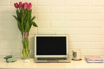 Beautiful and cozy place for a girl to work remotely.White table, laptop, vase with pink tulips, notebook and white porcelin cup. Office at home.