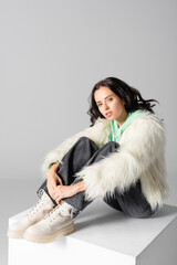  young woman in faux fur jacket posing on cube on white background