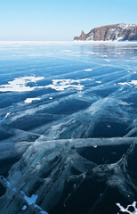 Beautiful winter landscape of frozen lake Baikal on a cold February sunny day. View from blue transparent ice with cracks to the famous Cape Hoboy on Olkhon Island. Ice travel. Natural background