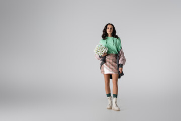  young woman in stylish winter outfit posing with flowers on grey background