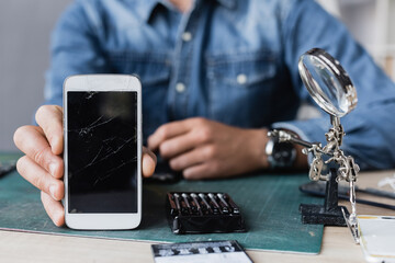 Cropped view of repairman showing smashed cellphone while sitting at table near magnifier and toolkit on blurred background