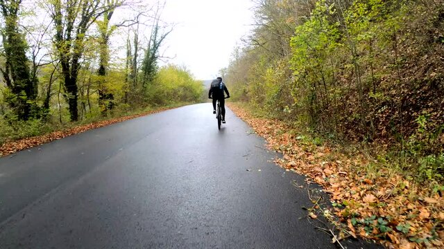 Cyclist riding a mountain bike on wet asphalt road through the autumn forest. Man in black clothes with a backpack cycling around nature.