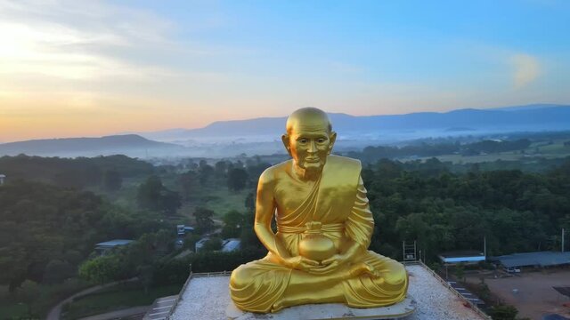 4k Aerial Big Luang Por Tuad monk statue surrounded by mountains of Khao Yai at sunrise in Thailand. Panning around of Luang Pu Thuat at dawn. Image of buddha disciple made of gold.