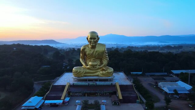 4k Revealing Aerial drone footage of a huge golden monk statue surrounded by mountains of Khao Yai at dusk in Thailand. Panning backward and up above Luang Pu Thuat image at dawn. drone flies back