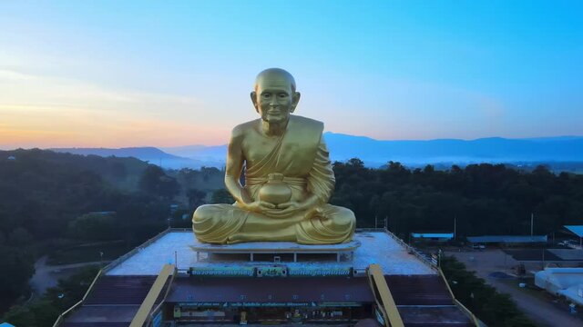 4k Aerial: drone flies forward to a huge monk statue surrounded by mountains of Khao Yai at dusk in Thailand. Panning forward to Luang Pu Thuat at dawn. Image of buddha disciple made of gold.