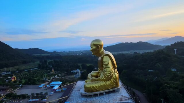4k 360 around Aerial drone footage of a huge golden monk statue surrounded by mountains of Khao Yai at dusk in Thailand. Panning backward and up above Luang Pu Thuat image at dawn. drone wraps around