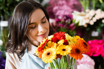 Fototapeta premium Brunette florist with closed eyes smelling gerberas in vase near blurred range of flowers on background