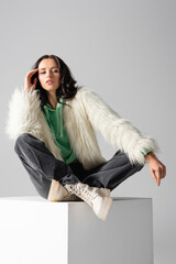  young woman in faux fur jacket posing on cube on white background